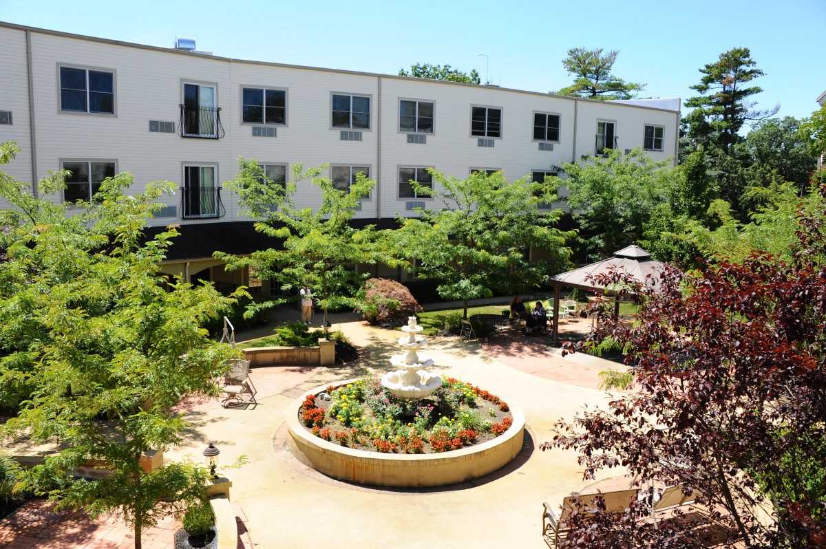 A serene courtyard featuring a central fountain surrounded by lush greenery and trees, adjacent to a multi-story building.
