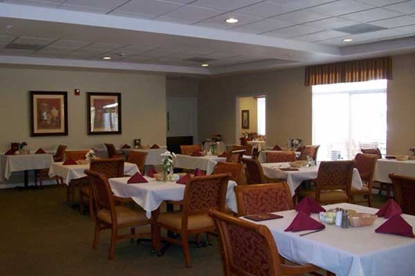 An empty dining room with neatly arranged tables and chairs, featuring white tablecloths and red napkins.