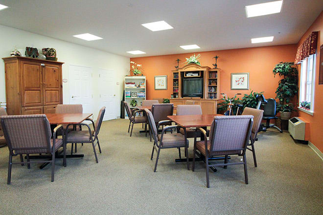 A community room with tables and chairs, featuring a television and bookshelf against an orange wall.