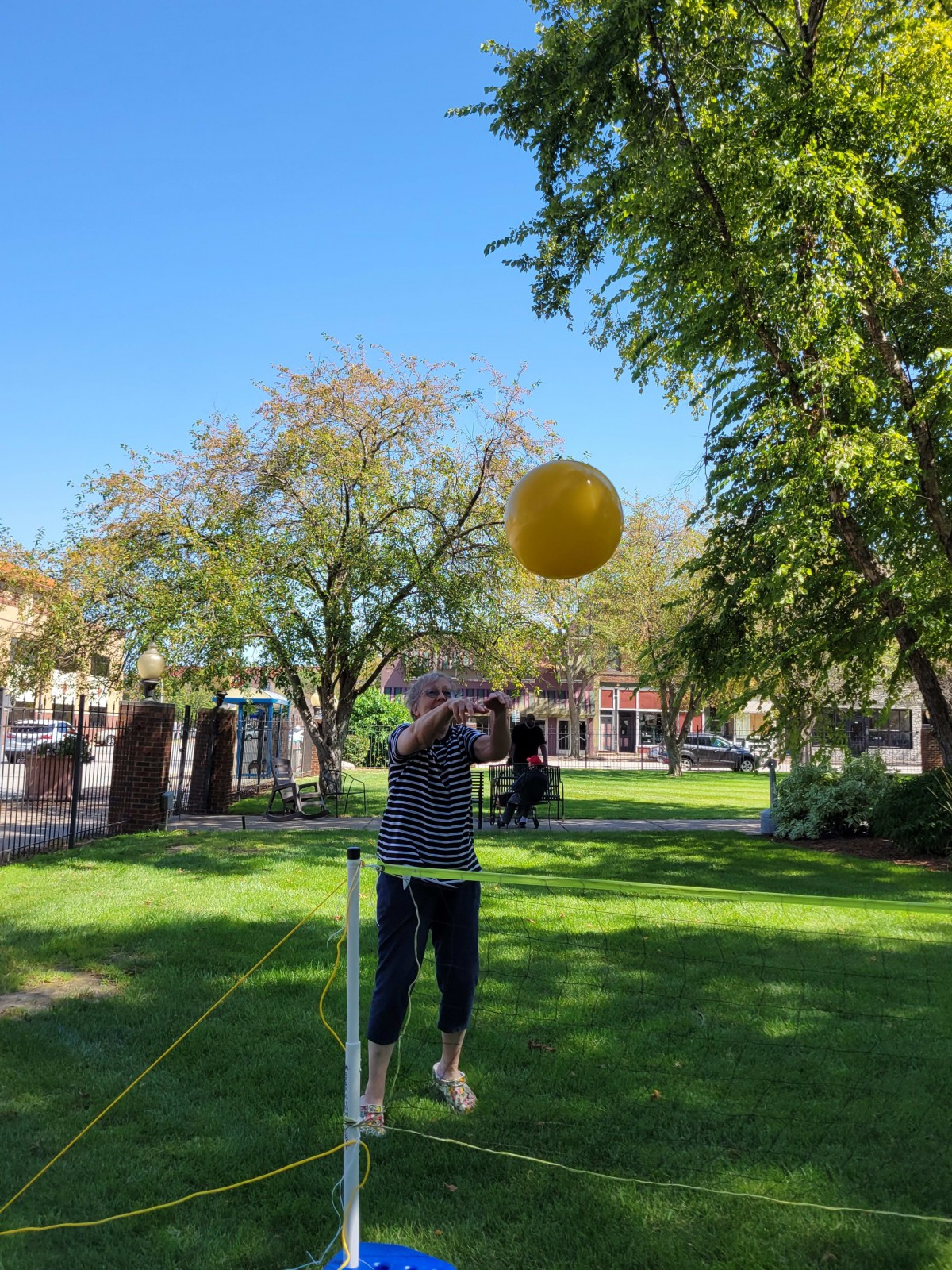 A person practicing balance on a slackline while reaching for a yellow ball in a sunny park.