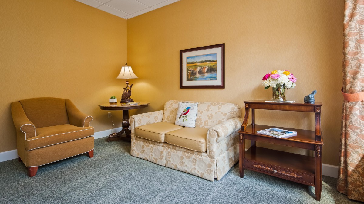 A cozy living room with a patterned sofa, armchair, side table with flowers, and a table lamp next to a piece of artwork on the wall.
