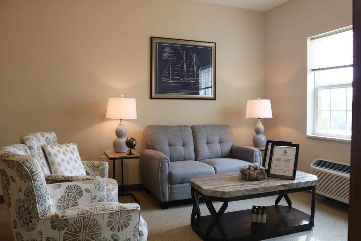 A cozy living room with a gray sofa, patterned armchair, wooden coffee table, and warm lighting from two lamps flanking the wall art.