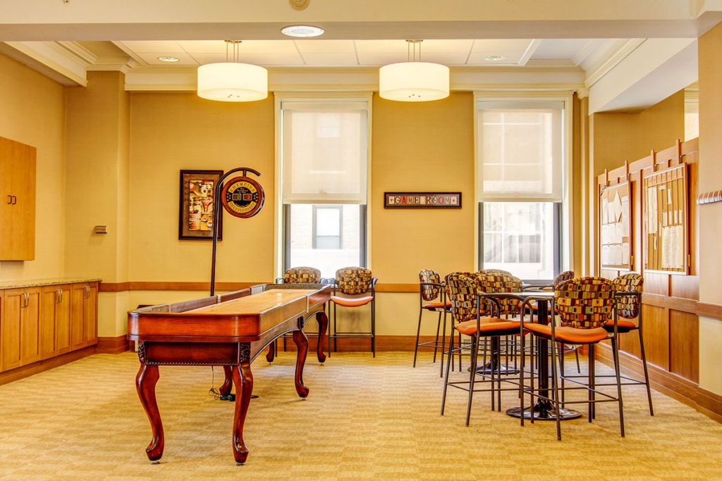 A cozy recreation room featuring a shuffleboard table, high chairs, and tables under soft lighting.