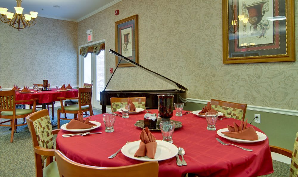 A dining room with a red tablecloth, set with glasses and napkins, featuring a piano in the corner.