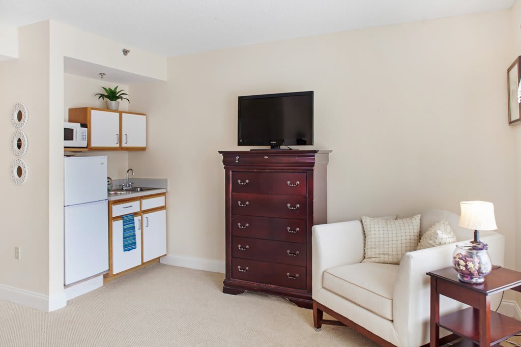 A compact living space featuring a kitchenette, a tall wooden cabinet with a television on top, and a comfortable chair.