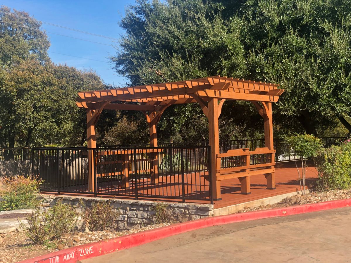 A wooden pergola with benches and a fence, set in a landscaped area with trees.