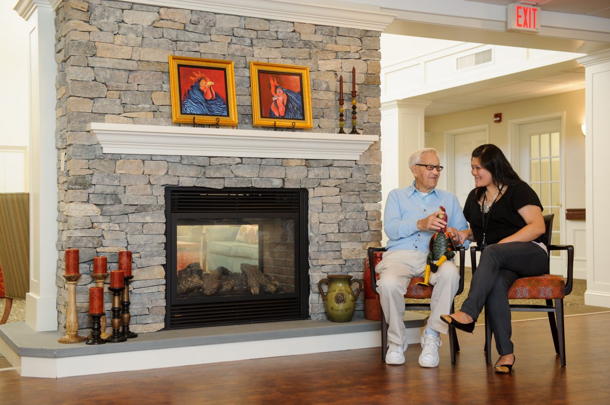 Two people seated in a cozy living room, engaged in conversation by the fireplace with portraits above it.