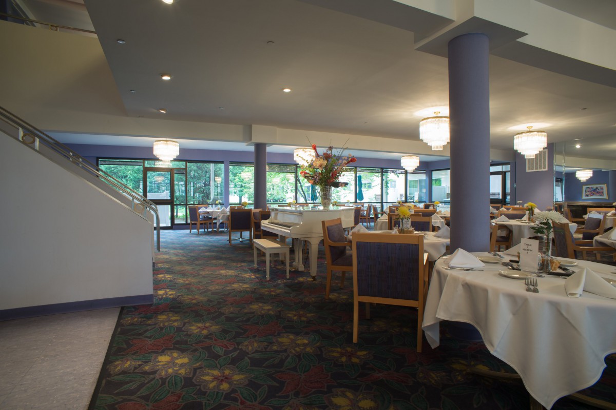 A spacious restaurant dining area with elegantly set tables and chairs, featuring large windows and chandeliers.