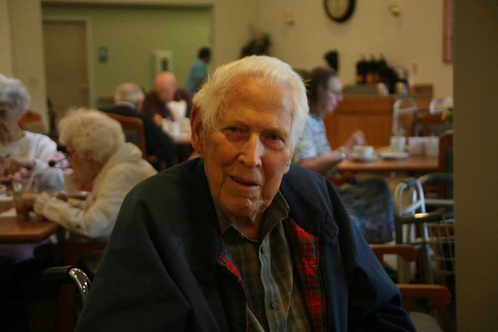 An elderly man sitting in a cafe setting with other patrons in the background.