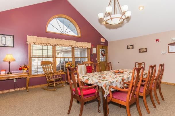 A cozy dining room with a large table set for a meal, adorned with a patterned tablecloth, surrounded by wooden chairs. The room features a vaulted ceiling with an elegant chandelier and large windows allowing natural light to fill the space.