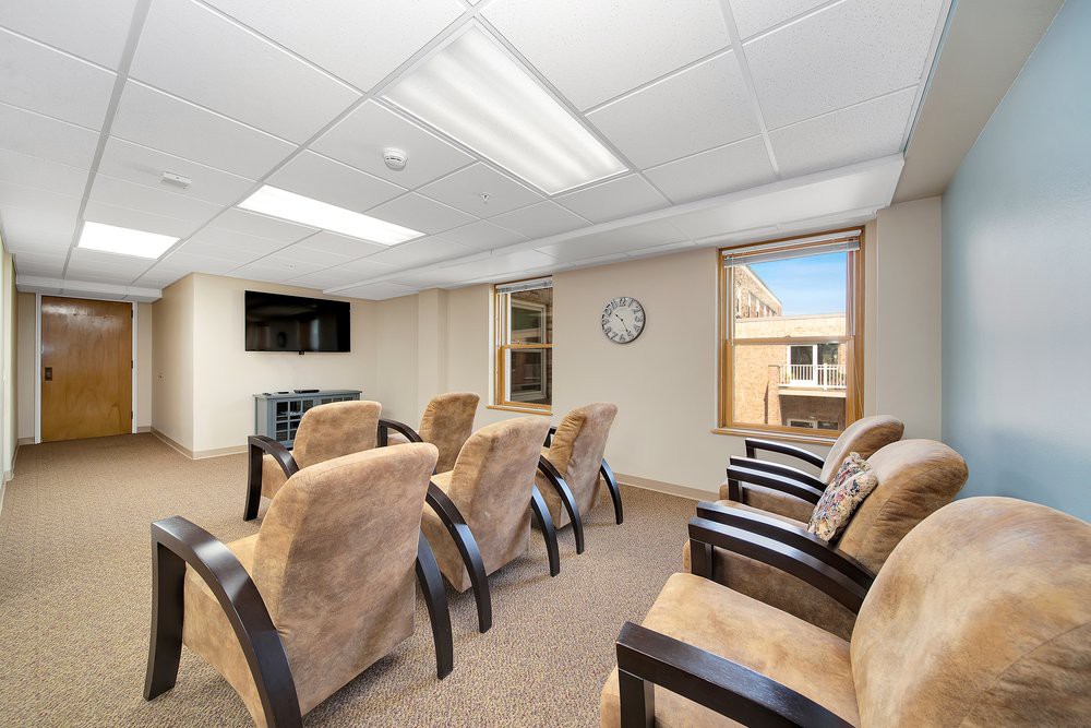 A small conference room with comfortable seating and a television, featuring large windows and ceiling lights.