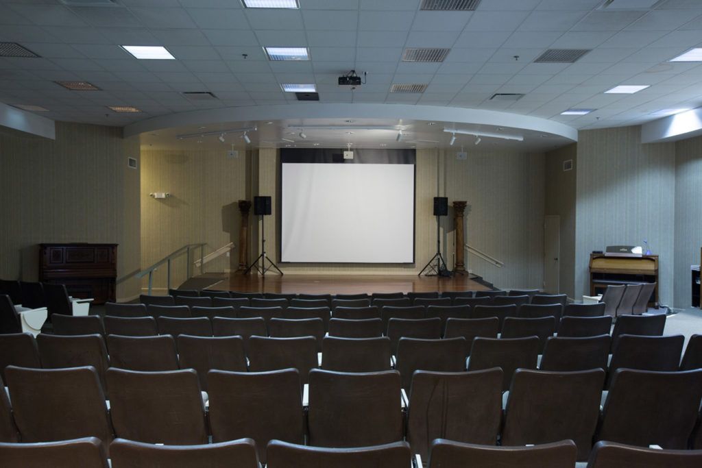 An empty auditorium with rows of seats facing a large projector screen on stage.