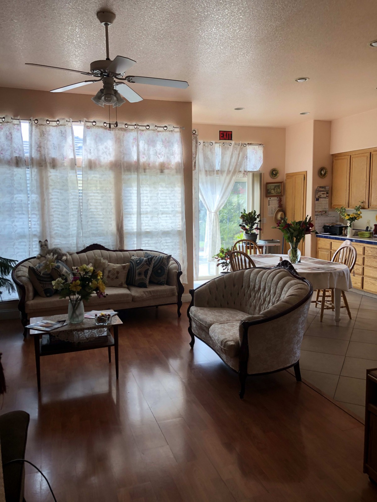 A cozy living room and kitchen area with an open floor plan, featuring wooden flooring and sunlight streaming through large windows.