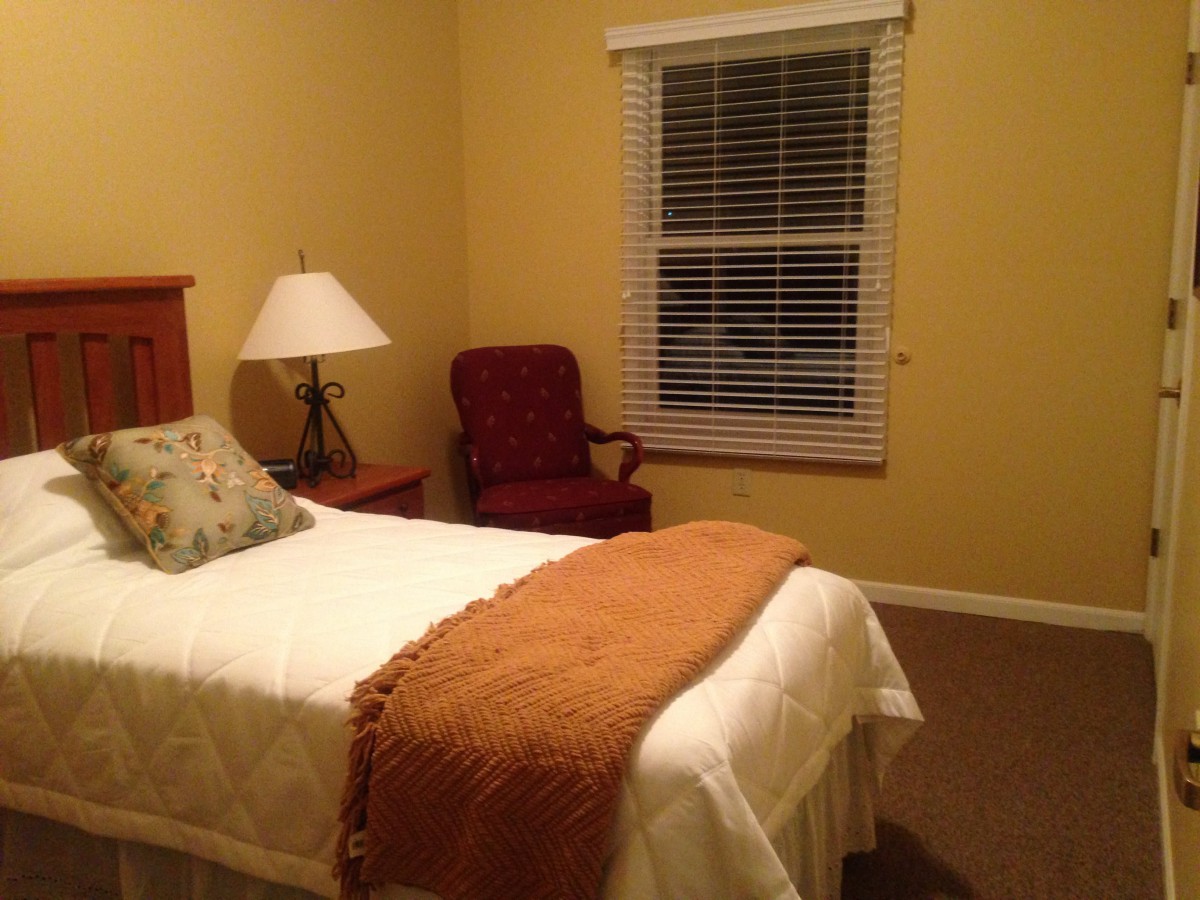 A cozy bedroom with a single bed, a lamp on a nightstand, an armchair, and a window with blinds.