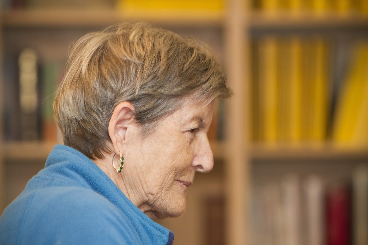 An elderly woman in profile view sits in front of a bookshelf.