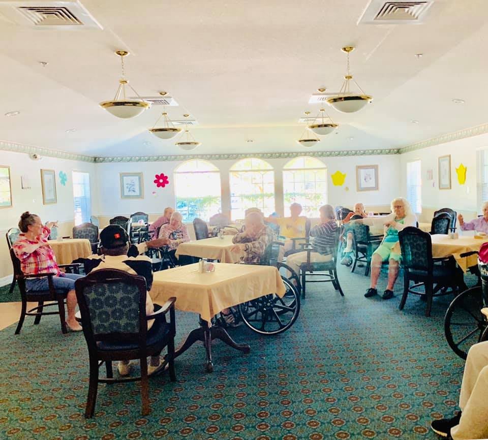 A bright dining room with elderly individuals seated at tables, some in wheelchairs.