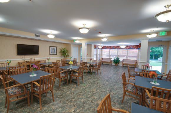 A spacious community dining area with wooden chairs and tables, decorated with plants and well-lit by ceiling lights.