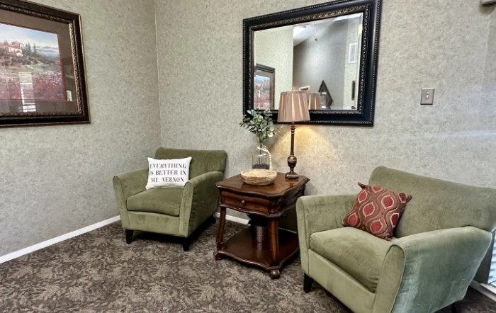 A cozy living room corner featuring green armchairs, a side table with a lamp, and a large mirror.