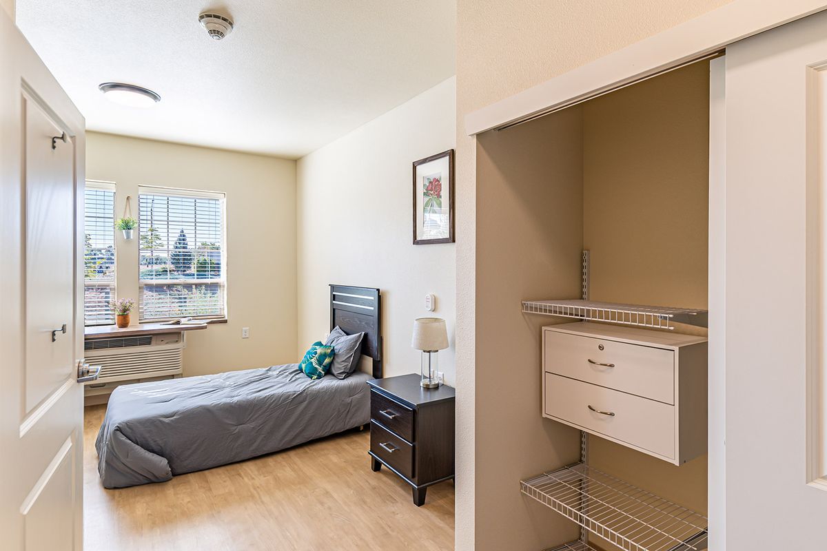 A minimalist bedroom with a single bed, nightstand, and an open closet, featuring wood flooring and natural light from the window.