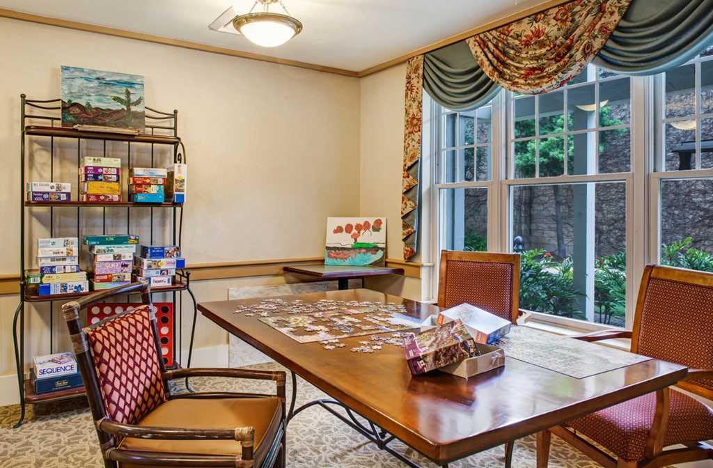 A cozy office space featuring a wooden table with a puzzle in progress, surrounded by chairs and illuminated by natural light from the window.