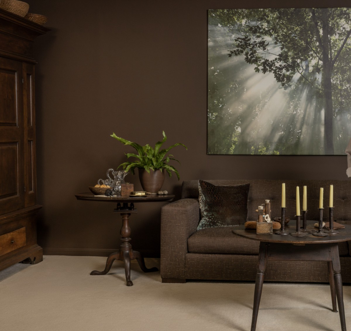 A cozy living room with brown tones, featuring wooden furniture, a sofa, decorative plants, and candles.