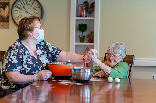 A caregiver and an elderly woman joyfully preparing a meal together at a dining table.