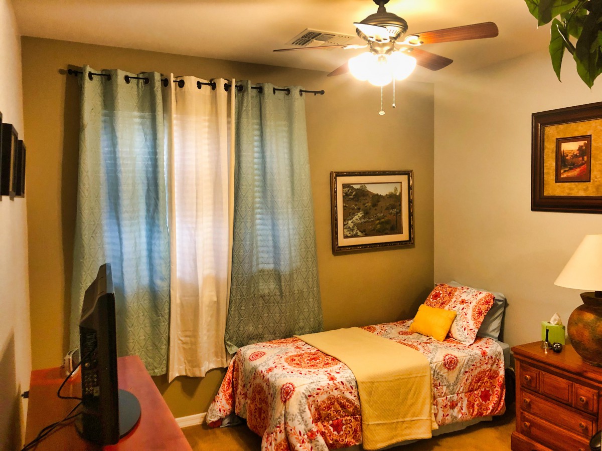 A cozy bedroom featuring a single bed with colorful bedding, curtains, a ceiling fan, and wall art.