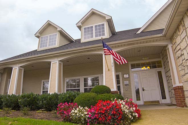 A well-maintained suburban house with an American flag displayed in the front yard, surrounded by manicured landscaping and vibrant flowers.