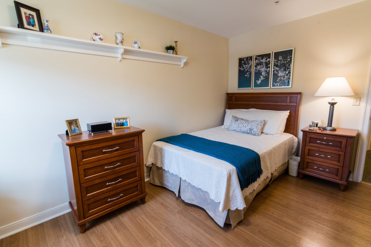 A cozy bedroom featuring a neatly made bed with a blue throw blanket, wooden dresser, and wall-mounted shelves adorned with small decorative items.