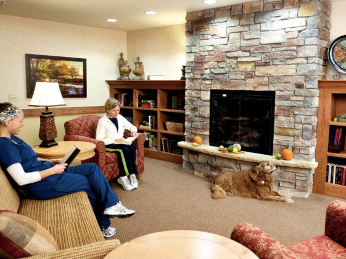A cozy living room with a stone fireplace, where two people are relaxing and reading near a bookshelf.