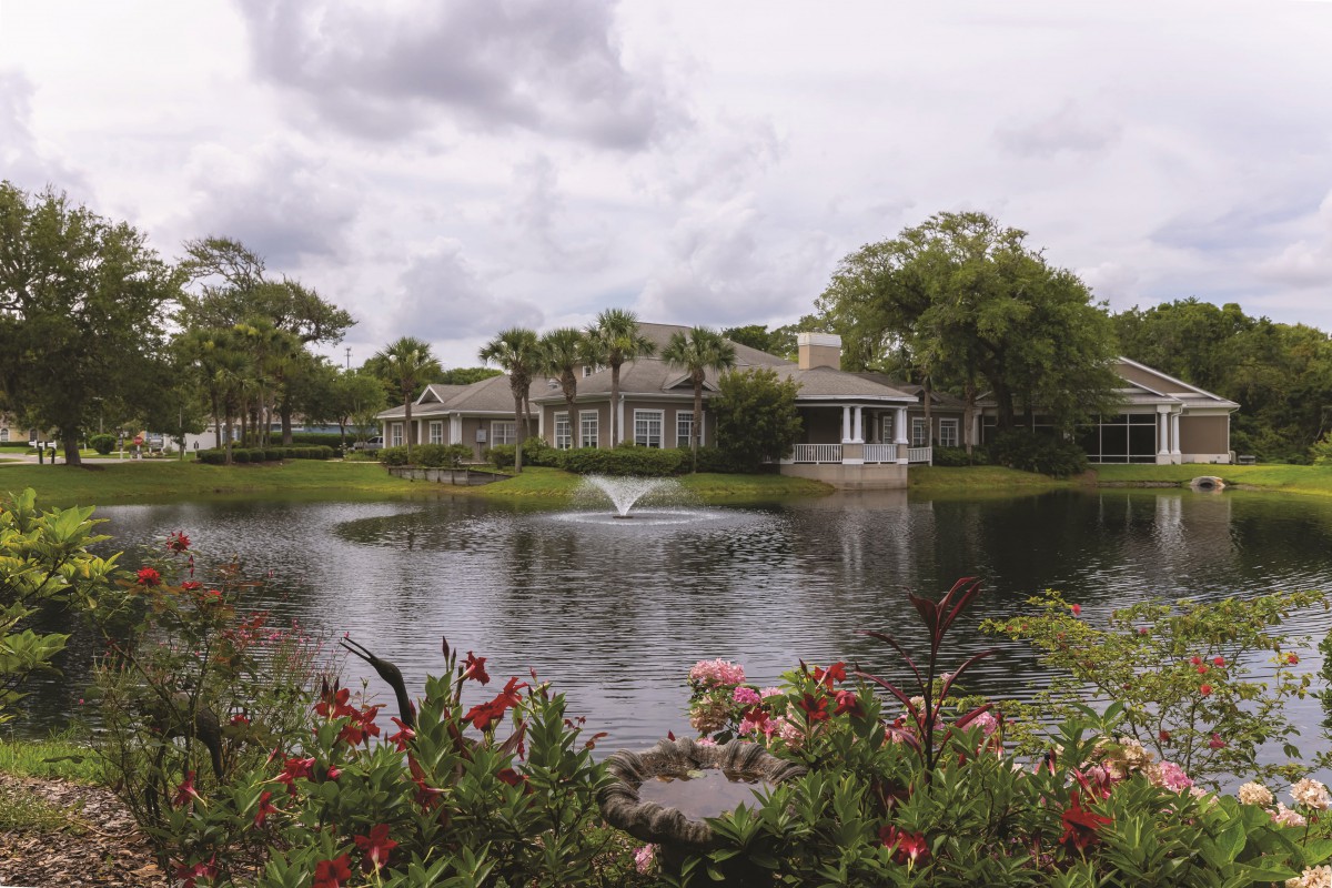 A beautiful house by a serene lake with a fountain, surrounded by lush greenery and colorful flowers under an overcast sky.
