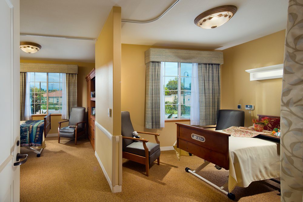 A well-lit hospital room with a bed, chairs, and large windows.