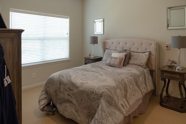 A cozy bedroom with a neatly made bed, a bedside lamp, and a window with blinds.