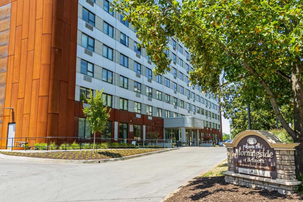 A view of a multi-story apartment building with a tree-lined road and sign in front.