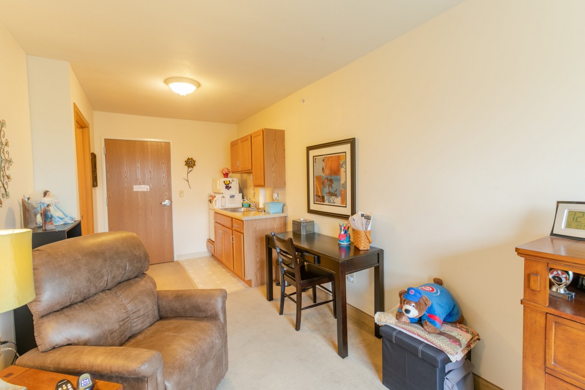 A cozy apartment living area featuring a sofa, small kitchenette, and a table with artwork on the wall.