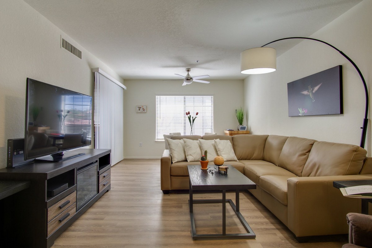 A modern living room featuring a comfortable L-shaped sofa, a flat-screen TV, and a stylish coffee table beneath a ceiling fan and large window.