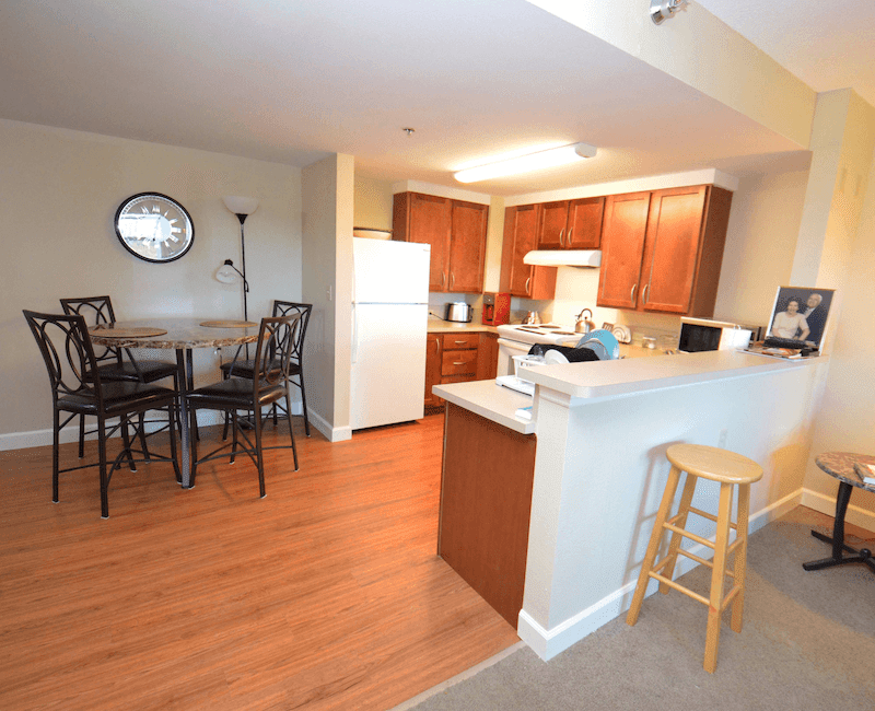 A modern apartment kitchen with dining area, featuring wooden cabinets and flooring.