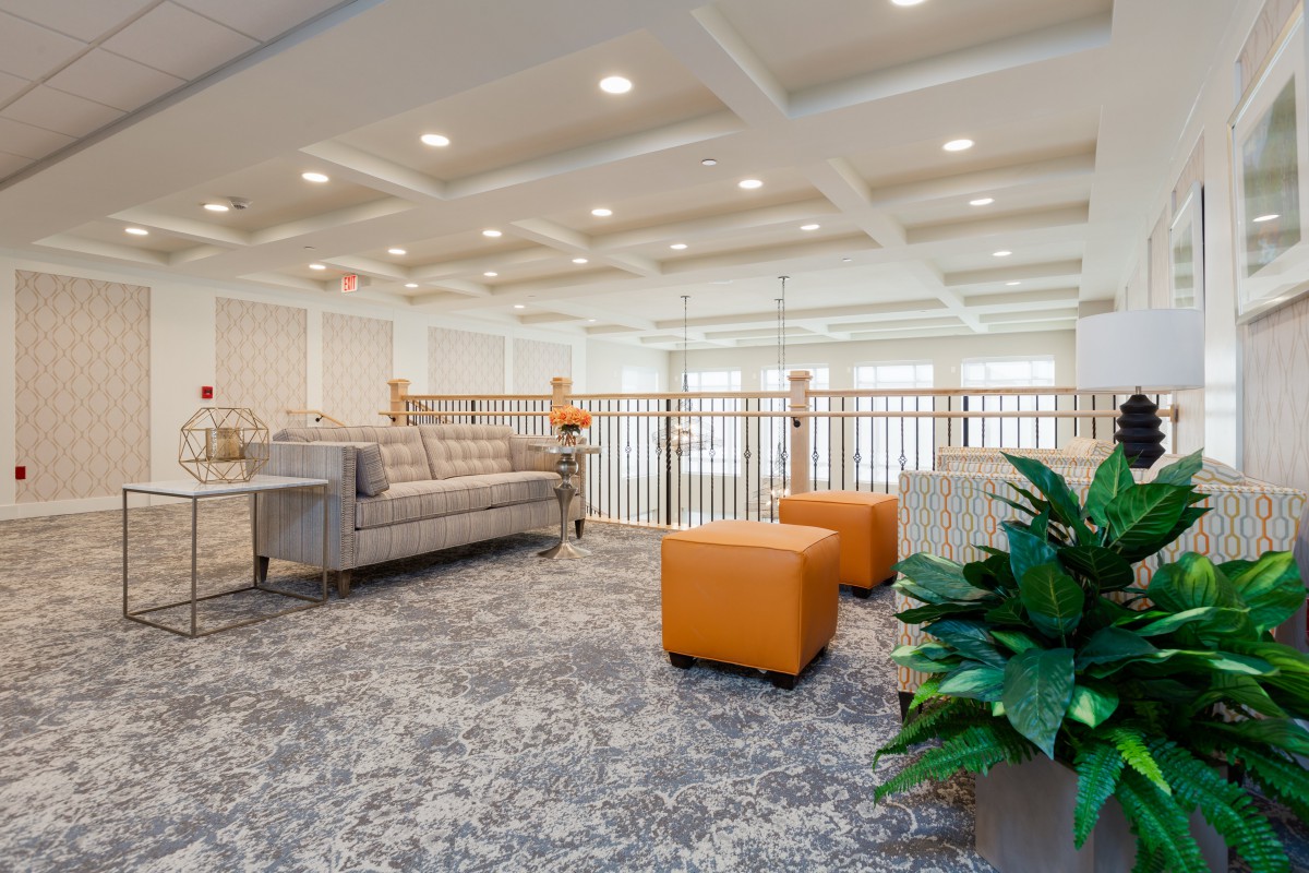 A modern lobby area featuring comfortable seating, including a sofa and orange ottomans, with decorative plants and stylish carpeting.
