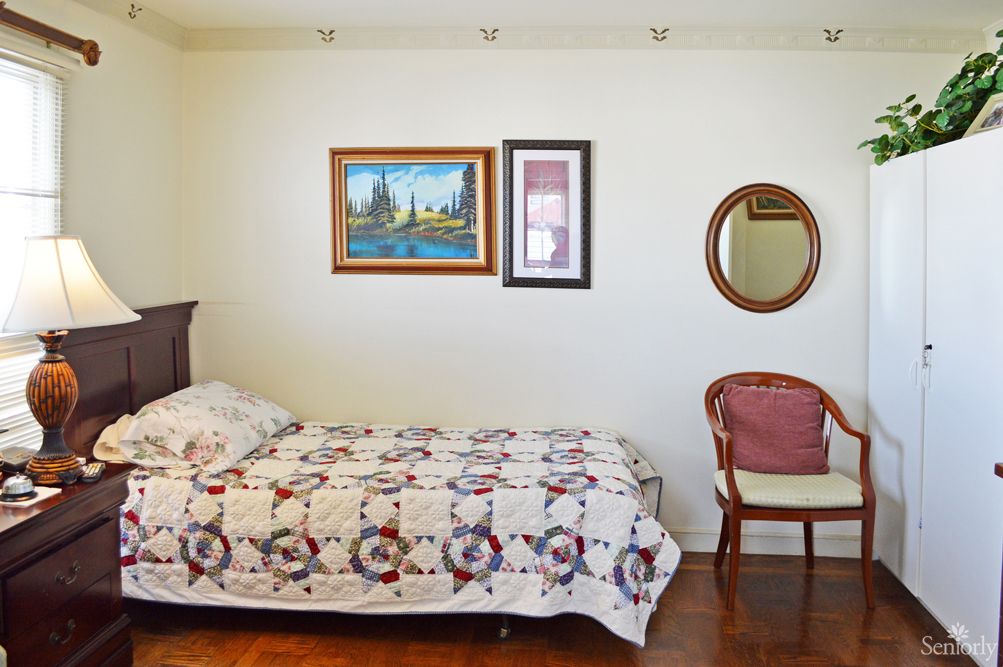 A simple bedroom with a single bed, decorated with a quilt, alongside paintings and a mirror on the wall, and a chair in the corner.