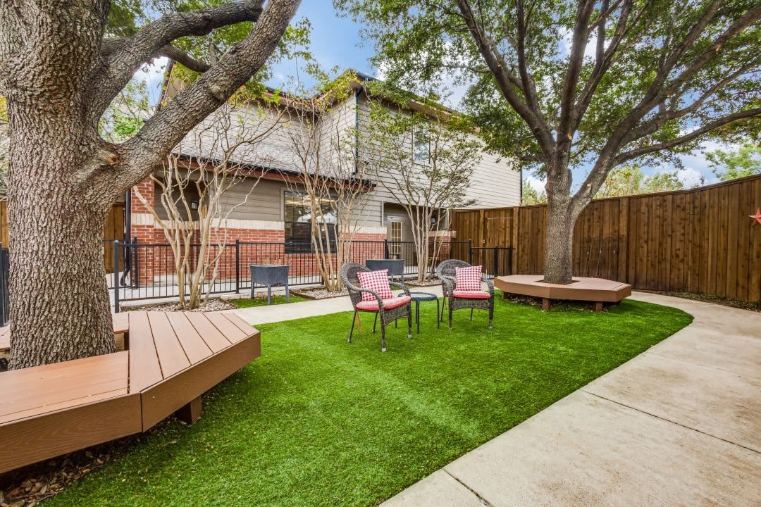 A cozy backyard with a lush green lawn, surrounded by trees and featuring several patio chairs and wooden benches.