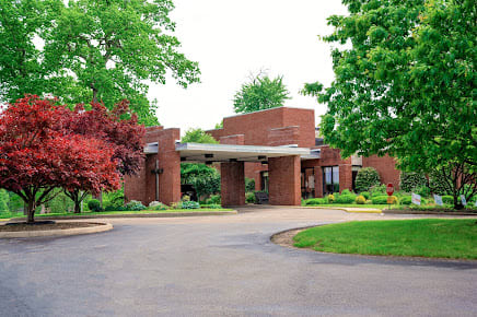 A red brick building surrounded by lush greenery and trees, featuring a curved driveway.