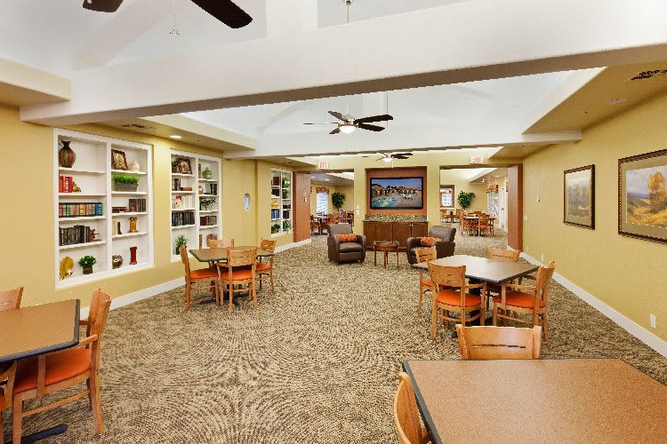 A spacious library room with tables and chairs arranged for reading or studying, featuring ceiling fans and bookshelves.