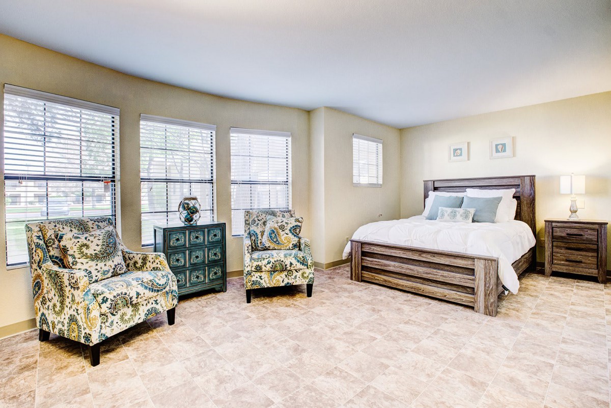 A cozy bedroom with a large bed, floral armchairs, and ample natural light.