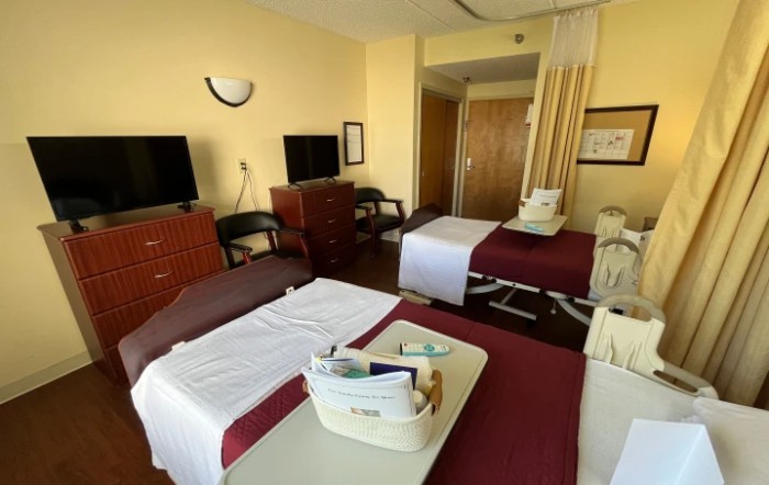 A hospital room with two beds, each equipped with tray tables and medical supplies. The room features a curtain divider, televisions, and chairs.