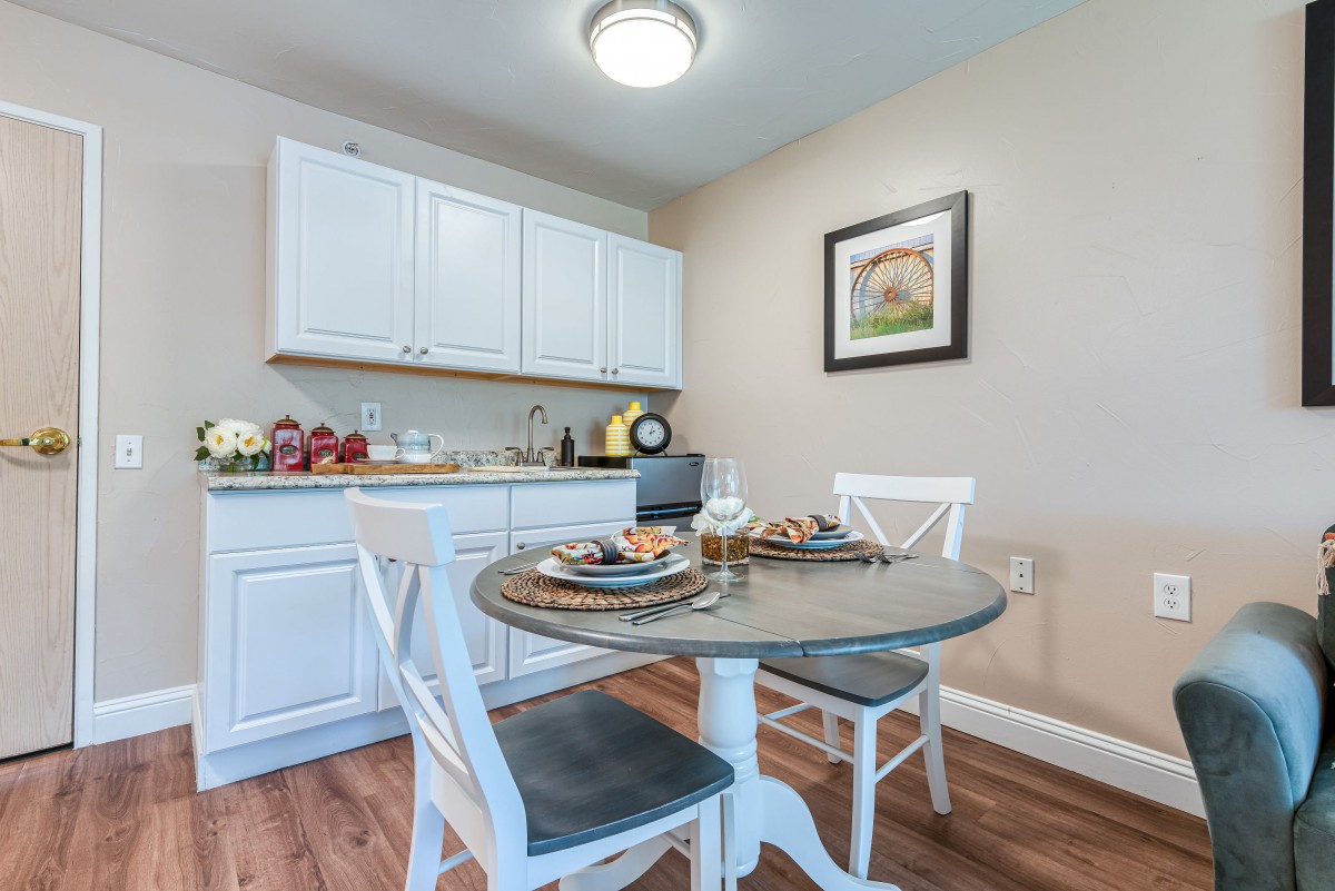 A cozy kitchen dining area featuring a small round table with two white chairs, surrounded by white cabinets and adorned with kitchen decor and artwork.