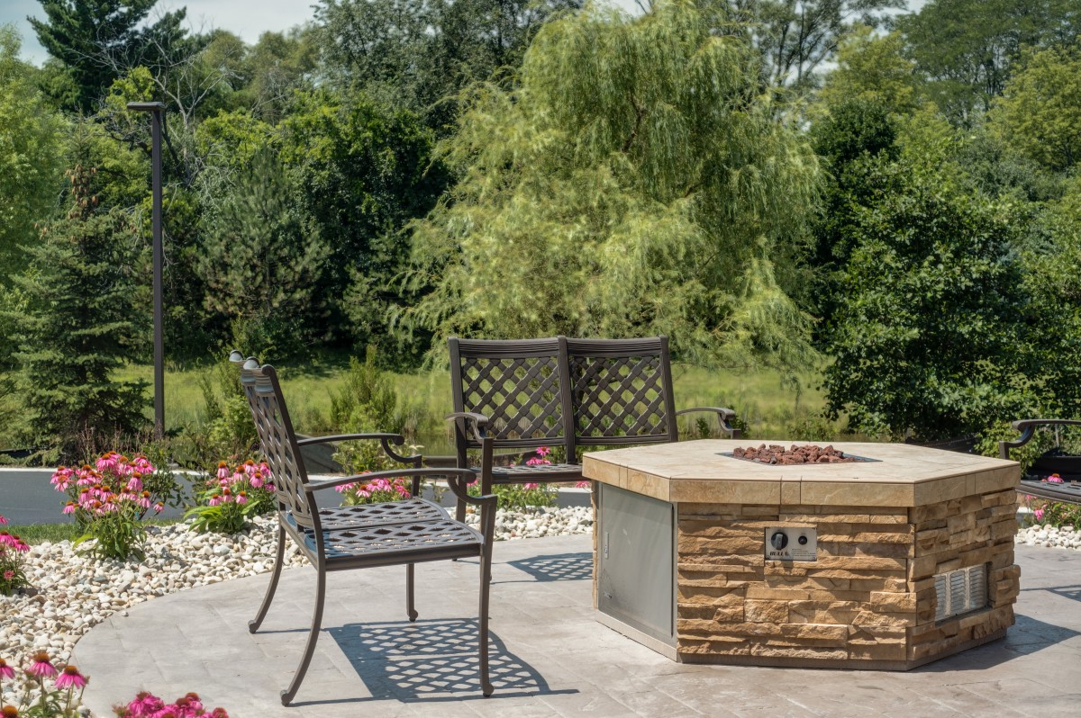A serene patio setting with a stone fire pit and metal chairs, surrounded by lush greenery.