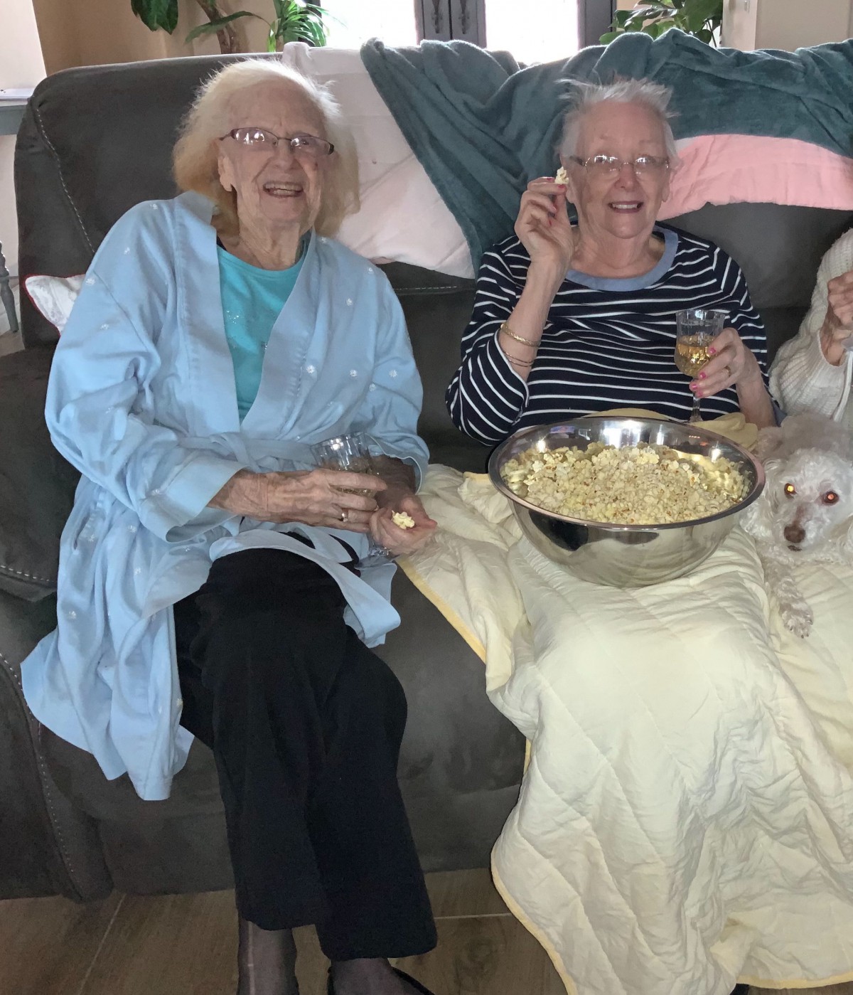 Two elderly women relaxing on a sofa, sharing popcorn with a small dog beside them.