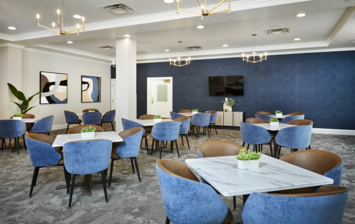 A modern dining area with blue chairs and white tables, featuring elegant chandeliers and a TV screen on the wall.