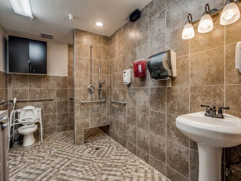A modern, accessible bathroom with tiled walls and floor, featuring handrails, a sink, and dispensers.
