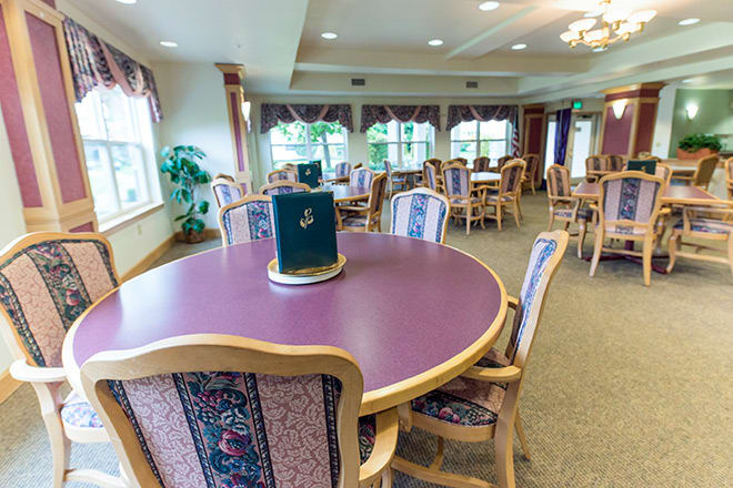 A well-lit dining area with multiple tables and chairs, featuring large windows and carpeted floors.